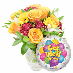 Presented on a pristine white background, this cheerful 'Get Well Soon' ensemble combines classic florist craftsmanship with playful celebration. The arrangement is positioned slightly to the left of centre in a clear glass vase so the stems and fresh water are visible, giving a sense of honesty and freshness. Several full, open roses in glowing orange and deep buttery yellow form the bouquet's focal point; their soft, velveteen petals exude a gentle, citrus-tinged fragrance that feels restorative. Bright yellow gerbera daisies punctuate the composition with sunny faces, while deep maroon or burgundy carnations weave texture and shadow between the blooms. Small clusters of white alstroemeria (Peruvian lilies) thread through the massed flowers, introducing delicate spears of light and a whisper of sweetness. Lush green foliage - glossy, varied, and supportive - frames the flowers, creating movement and a garden-fresh silhouette. Nestled to the lower right and partly overlapping the bouquet is a glossy silver Mylar balloon: its reflective field showcases a cheerful bunch of illustrated balloons in blue, purple, green, pink and yellow, some patterned with stripes, dots or checkerboards. The central message, Get Well Soon, appears in friendly yellow and blue lettering with a soft shadow, and playful starbursts dot the white border; a tiny KALEIDOSCOPE mark is visible at the edge. The image reads as a warm, optimistic gesture - exactly the sort of comforting gift someone in Golders Green might send to lift a neighbour's spirits during recovery.