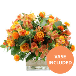 A rich, glowing floral arrangement in warm sunset tones is shown against a pure white backdrop, evoking the gentle light you might see over Golders Green at the end of a bright day. Dominating the composition are full, velvety orange roses, several with petals that blend into a softer yellow near their centres, creating a subtle ombré effect that draws the eye inward. Mixed among them are cheerful yellow garden roses, their petals tightly layered and slightly ruffled, adding a sense of abundance and comfort. Slender stems of peach and light orange alstroemeria weave between the roses, each Peruvian lily displaying fine speckles and elongated streaks on its inner petals, like delicate hand-painted details. Tucked thoughtfully throughout the bouquet are small, spherical, spiky orange accent flowers, reminiscent of safflower or thistle, which add a touch of playful texture and help the composition feel lively rather than formal. Surrounding the blooms is a generous halo of fresh greenery: softly rounded eucalyptus leaves in a silvery green tone mingle with deeper, glossy foliage, building depth and framing the brighter colours. All of the stems gather into a modern, square glass vase that reveals the clean lines of the arrangement and the clear water within, emphasising freshness and care. Around the vase's centre, a natural burlap or jute band introduces a rustic, tactile contrast that would look at home on a dining table near Golders Green station or in a living room off Hoop Lane. In the bottom right corner, a vivid orange circle carries the message "VASE INCLUDED" in bold white letters, indicating that this uplifting, seasonally toned bouquet arrives as a complete gift, ideal for celebrations, thank yous, or cheerful home décor.