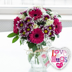 This image shows a lavish, contemporary bouquet that looks as though it has just been delivered to a cosy home off Finchley Road in Golders Green, bathed in gentle midday light. The flowers are arranged in a low, rounded clear glass vase, the stems carefully cut to an even length so they fan out in a perfect dome. Around the neck of the vase, a strand of natural twine is wrapped and tied, supporting a neat white gift tag printed with a stylised pink rose and friendly green script that suggests the blooms were thoughtfully chosen and gathered by hand. The arrangement is dominated by large gerbera daisies in a deep fuchsia-magenta shade, their petals radiating from dark, chocolatey centres, giving the bouquet a confident, joyful energy. Between these statement flowers, smaller two-toned chrysanthemums draw the eye: each bloom shows rich purple petals fading to white tips, circling bright yellow centres that gleam like tiny lanterns. Creamy white roses, some in full bud, others just opening, are tucked delicately among the bolder colours, offering a sense of calm balance and classic romance. Airy sprays of baby's breath drift through the design, their tiny white flowers creating a soft, cloud-like texture. Glossy green leaves and foliage frame the blooms, hinting at fresh, just-cut stems from a local florist's workbench. To the right of the vase floats a heart-shaped foil balloon, its metallic silver border and white interior catching subtle reflections. The balloon carries the playful message "Love n' Hugs 24-7" in a lively patchwork of pink, purple, and blue lettering, surrounded by small hearts, underlining the arrangement's purpose as a gift of constant affection. The background is a softly blurred, bright interior, suggesting a windowed room somewhere in Golders Green, turning the whole scene into a snapshot of celebration, comfort, and everyday romance.
