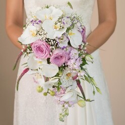 A romantic Golders Green bride stands lost in a tender moment, her arms embracing a lavish waterfall bouquet that embodies both classic beauty and modern finesse. Against the soft, muted tones of her surroundings-suggestive of a North London morning filled with potential-she holds her bouquet mid-torso, the draping florals capturing the delicate anticipation of a wedding day. The arrangement is a symphony of colours and textures: at its center, pristine white Phalaenopsis orchids with their golden hearts command admiration, surrounded by blush pink and lavender roses in varying stages of bloom. White sprays of baby's breath swirl like gentle mist between the more substantial blooms, their lightness balancing the composition. Trails of vibrant fuchsia Amaranthus flow downwards, intertwining with fresh, emerald foliage and unopened buds, adding dynamic movement and depth. Smaller purple blooms-perhaps reminiscent of Golders Green allotments bursting with summer stock-contribute layers of visual intrigue. The bride's gown, adorned with subtle lace, adds a sense of tradition and local legacy to the image. This bouquet, meticulously pieced together by Flowers Golders Green, combines emotive hues and natural forms to create an arrangement that resonates with the joy and elegance unique to wedding celebrations in the heart of NW11. The soft interplay of whites, lavish pinks, and serene lavender tones offers visual inspiration to couples in Golders Green planning a day filled with fresh beginnings, heartfelt promises, and floral artistry.