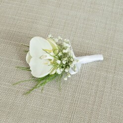 A refined white orchid boutonniere lies carefully arranged on a neutral, light beige fabric backdrop, whose subtle woven texture recalls the relaxed, understated interiors of cafes around Golders Green. At the centre sits a flawless Phalaenopsis orchid, its large, rounded petals an immaculate white that gently folds around a soft, creamy yellow throat, giving the flower a gentle depth and a serene, almost meditative presence. Encircling this focal bloom are airy sprays of white baby's breath, each tiny flower forming part of a mist-like cluster that creates a dreamy, romantic frame. Threaded through and beneath these delicate blossoms are slim strands of vivid green foliage, their fine, almost feathery leaves suggesting freshness and a quiet connection to nature, as if gathered from a shaded garden off Temple Fortune. The stems of the orchid, baby's breath, and greenery are drawn together with precision and wrapped in smooth white satin ribbon, the fabric folded snugly and finished with a modest bow near the top. The satin's faint sheen contrasts gently with the matte, organic textures of the flowers and fabric beneath, showcasing the careful hand of an experienced florist. Soft, even lighting washes over the scene, emphasising the clean whites and natural greens without harsh shadows, allowing the composition to feel calm and intentional. The boutonniere exudes classic wedding elegance, making it easy to imagine pinned to a groom's lapel as he steps into a synagogue near Golders Green Road or a registry office ceremony nearby. The overall impression is one of quiet sophistication, purity, and enduring love, perfect for formal events where subtle detail and craftsmanship truly matter.