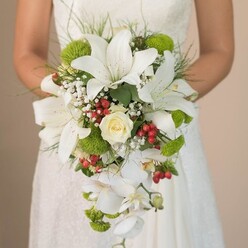 Cradled lovingly in the bride's hands, this cascading bouquet commands attention in the luminous ambiance of Golders Green. The focus falls instantly on show-stopping white lilies, their star-shaped blooms opening wide, each adorned with the faintest touch of burgundy speckles, drawing one's gaze inward. Nestled at the heart lies a classic, creamy rose, its tightly layered petals unfurling with soft promise-a floral centrepiece echoing the romance of a Finchley Road stroll. Alongside, trailing white Phalaenopsis orchids descend with stunning fluidity, creating a gentle waterfall that imparts an airy, sophisticated movement, as if inspired by the tree-lined roads near Golders Hill Park. Dotted among these elegant shapes, clusters of vivid lime-green cushion mums and buoyant tufts of gypsophila add joyful texture, while gleaming red Hypericum berries punctuate the bouquet, delivering bursts of colour that shine like autumn berries found in the local gardens. Fine wisps of grass-like foliage and dark leaves give the entire composition a natural, organic frame, balancing classic romance with contemporary verve. The bride's fingers, only just visible, suggest a quiet intimacy, her ivory gown a canvas for the bouquet's colours and form. Soft, diffused light enhances every textile nuance-the sheen of petals, the airy softness of baby's breath, and the tactile play of greens and reds. This bouquet, crafted with Golders Green's fresh florals, is an ideal muse for couples dreaming of a wedding rich with elegance, modern flair, and the welcome warmth of local tradition.