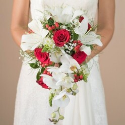 A vision of elegance from the heart of Golders Green, this bridal bouquet radiates romance and celebration, embraced delicately by the bride in her lace-adorned white wedding gown. Captured in rich, afternoon light, the bouquet's teardrop cascade features lush white oriental lilies, their intricately veined petals open wide, exuding sophistication and calm. Among them, soft white orchids trail gracefully downward, their gentle curves suggesting the serene flow of water in local Brent Park. Bold red roses, velvety and full, create a passionate contrast, their rich crimson tones echoing the spirit of new beginnings. Speckles of tiny white gypsophila and bright red hypericum berries are thoughtfully arranged, adding layers of lightness and playful texture, reminiscent of market blooms along Finchley Road. The foliage-a tapestry of feathery fern and deep green accents-frames the flowers, providing a touch of wooded tranquility known to the area. The bride's bare arms, softly freckles, offer a sense of intimacy, holding the bouquet close to her torso in a moment of joyful anticipation. The background's warm beige softness puts all eyes on the bouquet's artistry and florals, highlighting the expertise of Golders Green's florists. This bouquet captures the essence of commitment and style for weddings across NW11, perfectly suited to memorable ceremonies and warm gatherings in Golders Green.