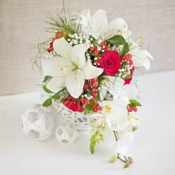 A luxurious floral centerpiece in crisp whites and deep reds commands attention on a pristine white surface, styled with the quiet confidence of an experienced Golders Green florist. The arrangement is presented in a round, open-work white basket that resembles delicately woven rattan, giving the display a handcrafted, artisanal feel. From this textured container rise generous white oriental lilies, their large petals elegantly arched and their golden stamens dusted with pollen that hints at a subtle, sweet fragrance. Interlaced among the lilies are plush red roses, some in tight buds that suggest new romance, others partially open with velvety petals spiralling around hidden hearts. Wisps of white gypsophila, or baby's breath, appear between the larger blooms, adding a fine, cloud-like softness that mimics lace veil detailing often seen in bridal fashion. Glossy red hypericum berries cluster here and there, bringing small accents of rich colour that feel particularly fitting for winter celebrations or candlelit dinners along Finchley Road. Framing the right side, a graceful trail of white phalaenopsis orchids spills outward, their arching stems dotted with open blooms and small green buds, each flower showing a delicate yellow and blush centre that enhances the sense of movement and luxury. Around the base, a trio of white woven spheres echo the basket's texture, tying the composition together like carefully chosen table decor at a wedding reception. Set against a softly textured beige backdrop, the whole scene feels serene, romantic, and meticulously styled, ideal for a wedding table arrangement, engagement celebration, or intimate family gathering in and around Golders Green.