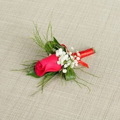 A romantic boutonnière is displayed on a neutral, pale beige background that looks like finely woven linen, its subtle texture echoing the calm, understated elegance of a North West London townhouse near Golders Green station. At the centre of the composition sits a single, tightly furled rosebud in vivid fuchsia pink, the petals smooth and satiny, curving inward in a compact spiral that hints at fullness yet to come. The colour is intense and jewel-like, somewhere between deep pink and magenta, and it glows warmly against the soft, natural backdrop. Clusters of tiny white baby's breath flowers nestle gently around the rose, their many minuscule blossoms creating a light, airy cloud that softens the bold central bloom, much like a veil softens a bride's features. Fine, feathery fronds of bright green foliage spread out from behind the rose, giving the boutonnière a graceful, slightly wild outline, as though it has just been gathered from a carefully tended garden off Finchley Road. These light green sprigs contrast with the deeper shadow at the base of the arrangement, where the stems are gathered and bound in a smooth, narrow strip of glossy red satin ribbon. The ribbon is wrapped with precision, forming a compact handle that adds a subtle glint of light whenever it catches the brightness of the scene. Illuminated from above, every detail is clear-the tiny petals of the gypsophila, the veins in the rosebud, the airy spaces between the greenery-conveying freshness, craftsmanship, and a sense of anticipation. This is the kind of lapel flower a groom, groomsman, or stylish guest might choose for a Golders Green wedding, a synagogue celebration, or a formal event where small details are thoughtfully considered.