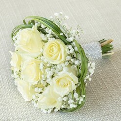 Presented atop a subtly woven pale beige fabric, this stunning bridal bouquet showcases the thoughtful artistry of a skilled florist in Golders Green. An array of eight to ten rich cream and ivory roses, varying from tight buds to fully open blossoms, radiates a serene luminosity, each petal softly curling outward as if to greet the day. Interspersed among these central blooms, lavish clusters of delicate white baby's breath enfold the roses like morning mist clinging to the fields near Golders Green's leafy Temple Fortune. Slender, bright green shoots-likely bear grass-are carefully looped throughout, their arcs imparting an energising, contemporary elegance to this otherwise classic arrangement. The bouquet's stems are aligned closely, expertly fastened in a luxurious silver-grey ribbon, meticulously detailed with symmetrical rows of gleaming pearls. This tactile handle feels cool and sophisticated to the touch while the fresh, natural ends of the stems evoke the vibrant heart of a market morning along Finchley Road. Such a bouquet, ideal for a celebratory wedding or a memorable anniversary in NW11, blends refinement with organic beauty, perfectly encapsulating the quiet sophistication of Golders Green.
