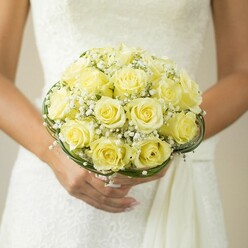 Captured in the serene ambiance of a Golders Green wedding, this bridal bouquet radiates refined beauty and heartfelt sentiment. The arrangement, seen cradled lovingly by a bride above her intricately detailed gown, showcases an array of pale yellow roses, velvety and fresh, each blossoming in a soft spiral that mirrors the tender joy of the occasion. Sprigs of baby's breath float delicately among the roses, their tiny, airy white blooms imparting a dreamlike quality reminiscent of early morning mist rolling over local gardens near Hoop Lane. Tucked below, graceful arcs of deep green leaves or decorative wire echo the gentle curves of the bouquet, lending both structure and artistry known to Golders Green's expert florists. The bride's hands-elegantly manicured-frame the flowers with gentle care, her commitment symbolized by a shining ring as sunlight plays across the surface of the petals. The softly blurred background, likely a tastefully appointed venue in the neighbourhood, keeps attention centered on the luminous bouquet and the rich texture of the gown. Every element speaks of understated celebration and hope, making this photograph ideal for wedding inspiration or for couples envisioning an elegant ceremony in the Golders Green area, accompanied by timeless floral artistry.