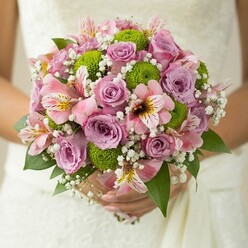 Under the gentle light of a Golders Green morning, a bride's hands cradle an exquisite bouquet-each element thoughtfully chosen for its beauty and meaning. Plush roses in charming shades of lavender and blush pink provide a timeless base, their aroma subtle but evocative. Nestled amongst them, lively lime green button chrysanthemums punctuate the design with their spherical, playful forms, introducing a spark of freshness to classic romance. Pink alstroemeria lilies, with their intricate speckled hearts of yellow and maroon, add interest and complexity, reminiscent of the vibrant diversity found along Finchley Road. Wisps of baby's breath, delicate as early mist hovering over Golders Hill, are tucked with care between the main blooms, lending an airy softness to the bouquet. Glossy deep green leaves accentuate the pastel tones and provide natural structure. The composition is nestled against the soft folds of a lacy wedding gown, each detail tailored as lovingly as a bespoke arrangement from a neighbourhood florist. Whether destined for a wedding at a Golders Green synagogue or an intimate celebration nearby, this bouquet reflects both the artistry and heartfelt joy of local floral tradition-a fresh, textural creation to be remembered for years to come.