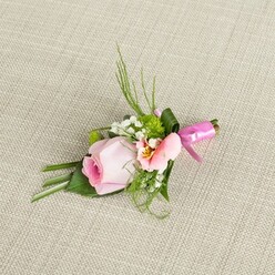 Evoking the understated charm of a spring morning in Golders Green, this floral boutonniere rests serenely upon a finely woven linen background, its neutral tones setting the stage for exquisite floral artistry. The centrepiece-a blush-pink rose, petals elegantly spiraled and promising newly opened beauty-anchors the arrangement. Alongside, a softly ruffled bloom in hues of pale peach and pink unfurls, brightened by golden undertones and gentle maroon flecking near its throat, inviting comparisons to the delicate borders seen along Hampstead Heath Extension nearby. Tiny, snow-white clusters of baby's breath nestle amid bold orbs of green cushion mums, weaving a subtle texture reminiscent of nature's careful balance. Wisps of deep emerald grass and fernlike fronds extend upwards, while wide, glossy leaves cradle the flowers, some artistically folded for a structured flourish, echoing the cultivated greenery of Golders Green's gardens. The stems are gathered gently and secured with a vibrant fuchsia ribbon, expertly tied into a fine satin bow, adding a celebratory accent that captures light and attention alike. Diffused natural lighting highlights the gently dewy petals and crisp foliage against the softly coarse, oatmeal-hued textile, bringing out every layer of detail and artistry. This graceful accessory-crafted for special occasions from weddings at local synagogues to proms and milestone celebrations-captures the spirit of community and refinement that defines Golders Green, a floral keepsake ready to mark cherished moments with elegance and style.