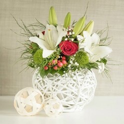 A sophisticated floral centerpiece sits quietly on a white surface, evoking the relaxed elegance of a Golders Green reception venue near Temple Fortune. At its heart is a distinctive round white vase, sculpted from interlacing bands that form an airy, open-weave texture, like a modern basket rendered in porcelain. From this sculptural container rises a lush bouquet in a classic white, red, and green palette. Several white lilies, some fully unfurled, others still held in sleek green buds, create sweeping lines and a sense of openness, their star-shaped faces and golden stamens radiating purity and calm. Tucked between them are deep red roses, their velvety petals folding inward in tight spirals that suggest romance and heartfelt emotion-perfect for a wedding table or engagement dinner. Bright green spherical blooms, like playful pom-poms of dianthus or chrysanthemum, punctuate the arrangement with vivid freshness. Sprays of baby's breath weave among the larger flowers, their tiny white blossoms adding a soft, mist-like layer. Clusters of red berries add warmth and a subtly festive accent, while cascading greenery-fine fronds mingled with darker, glossy leaves-spills over the edges, giving the bouquet a naturally abundant shape. To the left of the vase rest two decorative woven balls in pale, natural tones, mirroring the vase's pattern and adding a stylish finishing touch. Set against a gently textured light beige wall, the arrangement feels serene yet celebratory, ideal for wedding breakfast tables, anniversary dinners, or refined home entertaining in and around Golders Green, with the freshness and care you'd expect from a local North London florist.