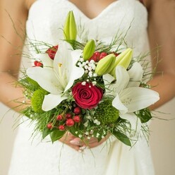 This close-up captures a moment of anticipation and joy as a bride in Golders Green cradles her elegantly designed bouquet, reflecting the artistry and romance woven into every wedding day. Her textured white dress forms the perfect canvas for an exuberant blend of flowers-a union of pure white oriental lilies, their wide petals unfolding gracefully, cradling golden stamens at their heart. Intertwined, deep red roses radiate passionate warmth, their classic spirals exuding time-honoured romance. The ensemble is further enlivened by unopened, vibrant green lily buds-a flourish of life and hope that resonates with the lush gardens dotting the Golders Green neighbourhood. Speckles of soft, snowy Gypsophila (baby's breath) impart a dreamy haze, evoking wishes whispered into the late afternoon air, while brilliant red hypericum berries scatter joyful colour reminiscent of a walk past Golders Green's thriving Saturday market. Spherical green button florals-perhaps green trick dianthus-nestle amidst the rich variety of foliage, from sleek emerald leaves to feathery ferns, building depth and a natural sense of abundance. The gentle grip of the bride's hands beneath the bouquet amplifies its meaning-precious, grounding, and full of hope. This bouquet, lovingly arranged by a local floral designer, is the epitome of heartfelt craftsmanship for a Golders Green ceremony, promising beauty and memory woven delicately together.