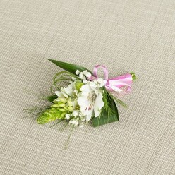 A compact and elegant floral boutonniere lies on a pale beige, linen-like fabric, the soft weave of the cloth forming a calm, neutral stage that enhances the crisp colours of the flowers. At the centre sits a white alstroemeria, sometimes known as a Peruvian lily, its petals wide and slightly ruffled at the edges, opening generously to show a throat flecked with fine reddish-brown streaks. These delicate markings, along with the visible stamens, give the flower a sense of character and maturity, as if it has just fully opened for the day's celebration. To one side rises a sculptural column of lime-green buds, tightly clustered in a conical form that suggests an Ornithogalum spike, adding interesting height and texture. Threaded among these focal elements are airy sprigs of baby's breath, each tiny white blossom like a dot of light scattered around the main bloom, lending a dreamy, romantic softness. Deep green, lance-shaped leaves cradle the composition, while more slender, grass-like strands curve around the edges, introducing gentle lines and an organic sense of movement, as though caught in a light breeze on Golders Green Road. At the right, a neatly tied bow in bright pink satin secures the stems, its smooth, reflective surface creating a cheerful pop of colour that hints at joy and celebration. The lighting is even and natural, showing off the fresh, firm petals and healthy foliage without harsh shadow. The overall mood is serene yet festive, the sort of carefully considered detail a couple might choose for a wedding in Golders Green or a formal event near Brent Cross, where a small piece of floral artistry can quietly complete a polished, personal look.