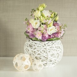Bathed in soft, diffused daylight, a thoughtfully arranged bouquet becomes the focal point-a masterful celebration of organic beauty and contemporary craft from the heart of Golders Green. The white vase, woven from painted rattan strands, is as much a piece of art as a vessel, its open structure giving a sense of airiness. Rising from this nest-like base, creamy white and pale yellow lisianthus blooms unfurl in gentle ripples, paired harmoniously with vibrant blush-pink stock that adds graceful height and movement. Just below, clusters of deep purple aster or allium nestle among glossy, dark green leaves, the whole arrangement forming a lush, cohesive tableau. Two decorative spheres, fashioned from light natural rattan with beautifully intricate patterns, rest confidently to the left, echoing the vase's modern, woven motif and reinforcing the handcrafted theme beloved by Golders Green's creative community. The entire composition rests on a gleaming white tabletop and stands before a muted greige wall-its subtle texture offering a sophisticated backdrop, much like the local architecture of NW11. An atmosphere of calm and understated celebration radiates from the arrangement, making it an ideal choice for elegant wedding receptions, intimate anniversary meals, or those seeking a touch of refined tranquility in their lives. With every stem carefully placed by a local florist, the piece captures a feeling of fresh beginnings and natural elegance that's at home on Golders Green's tree-lined avenues.