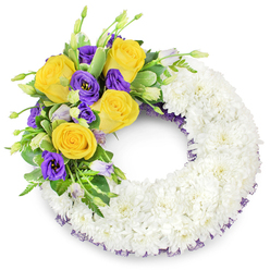 Presented on a clean white backdrop, this circular sympathy wreath is a study in texture and tonal restraint crafted for moments of remembrance in Golders Green. The foundation is a dense, continuous ring of pristine white spray chrysanthemums; their tiny petals are arranged so tightly the surface becomes an even, cloud-like band that speaks of serenity and collective comfort. A carefully composed accent occupies the upper-left quadrant: several large, honey-yellow roses fully unfurl their petals in soft spirals, offering a gentle warmth that contrasts with the cool, velvety presence of deep purple lisianthus (Eustoma). The lisianthus' ruffled cups and a few lighter lavender buds introduce depth and a muted lyrical quality, while small unopened buds and slender green stems suggest quiet renewal. Supporting foliage - glossy salal-like leaves, delicate fern fronds and a few variegated leaves - is woven through the cluster to create movement and a natural frame for the blooms. Peeking from beneath the chrysanthemum base along the inner and outer edges is a narrow, dark purple mesh ribbon that discreetly echoes the lisianthus tones and gives a subtle finished edge. In soft daylight the chrysanthemums read as pure and matte, the roses as satiny and fragrant, and the composition as composed and consoling. As florists serving Golders Green - including arrangements for services at local churches and the Crematorium - we at Flowers Golders Green design pieces like this to convey sympathy, to sit respectfully beside an open book of condolences, and to travel well for local collection or delivery when families need a dependable, compassionate tribute.