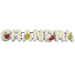 The image shows a heartfelt GRANDMA floral tribute laid out in large capital letters, each one carefully built from hundreds of fresh white blossoms. The letters appear to float against a bright white background, so the gentle shadows and rich textures of the flowers can be clearly sensed. Every letter is carpeted in tightly clustered white chrysanthemums or similar small blooms, giving the surface a plush, cloud-like softness that feels calm and reassuring. Just beneath the main blooms sits a narrow frill of light lavender or soft purple flowers, tracing the silhouette of each letter and adding subtle depth and contrast. On the lower curve of the opening 'G', three deep red roses glow like precious keepsakes, hugged by glossy green leaves that bring a natural woodland touch. The first 'A' carries two striking chartreuse orchids, their yellow-green petals and reddish-brown throats poised along the crossbar like delicate wings. The central letters 'R', 'N', and 'D' remain pure and uncluttered, their white faces edged with lavender, allowing the coloured highlights to guide the eye gently across the word. Near the end, the strong verticals of the 'M' cradle another tight group of red roses, echoing the love expressed at the beginning, while the final 'A' repeats the distinctive green orchids, tying the tribute together. All the letters are anchored to a discreet dark metal bar for stability, designed to sit proudly at a funeral service, wake, or graveside ceremony in and around Golders Green. The overall feeling is one of tender remembrance and gratitude for a grandmother's life, quietly evoking shared stories from home, local parks, and familiar streets.