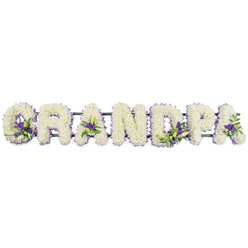 A lovingly crafted floral lettering tribute spells out GRANDPA in bold, upright capitals, aligned in a neat horizontal row as though placed along the front of a coffin or easel stand during a quiet service in Golders Green. Each letter is completely carpeted with compact, creamy white chrysanthemums or carnations, the tiny flower heads pressed closely together to form a smooth, cushion-like texture that looks both soft and substantial. The white surface suggests innocence, respect, and a peaceful goodbye, catching the light in gentle highlights and shadows that reveal every curve and edge of the word. Scattered with intention through this white canvas are small groupings of rich purple and lilac blossoms, perhaps lisianthus or short-stemmed chrysanthemums, partnered with glossy green leaves. You can see a concentrated cluster touching the lower curve of the 'G', another resting calmly in the open center of the first 'A', then further accents shaping the 'D', brightening the 'P', and settling into the final 'A'. These tonal contrasts add depth and a quiet sense of hope, like spring flowers glimpsed in a corner of Golders Green Crematorium's gardens. Beneath the flowers, a sturdy dark green frame links the letters together, almost invisible yet essential, so the name displays as one continuous tribute. The arrangement feels deeply personal, the sort of bespoke funeral design that Flowers Golders Green might prepare for a family wishing to honour a devoted grandfather, with careful attention to freshness, detail, and timely delivery to local services around NW11.