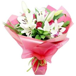 An opulent bouquet of white lilies and red roses is shown in close detail, photographed against a clean white background that makes every colour and texture stand out, much like a display in a boutique florist just off Golders Green Road. The central lilies are large Oriental blooms, their open, star-like petals a pristine, velvety white that curves around deep centres marked by clusters of reddish-brown stamens. Some blossoms are fully open, with petals sweeping outward in graceful arcs, while a few buds, tipped with pale green, remain closed, suggesting the bouquet will continue to evolve and open over several days. Threaded between the lilies are velvety red rosebuds in varying stages of bloom: some are tight, jewel-like spirals, while others are beginning to loosen, their layered petals revealing depth and richness that adds a sense of romance and warmth. Encircling the flowers is a generous collar of foliage, with deep green, glossy leaves creating a solid backdrop and finer, spiky strands of greenery adding movement and height, as if stirred by a gentle breeze on a walk through nearby Hampstead Garden Suburb. The entire composition is gathered into a voluminous wrap of bright pink, glossy cellophane, slightly translucent so that hints of stem and leaf show through the folds. The crinkled texture of the wrap catches the light in soft ripples, adding a celebratory sparkle. At the base, a slim, lime green ribbon is tied snugly around the stems, giving a crisp, fresh accent. The bouquet radiates thoughtful celebration-equally at home as a romantic gesture, a Mother's Day surprise, or a congratulatory delivery to a doorstep in Golders Green.