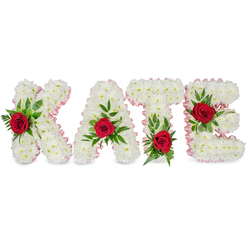 A hand-crafted memorial composition spells KATE in substantial, three-dimensional block letters, each letter formed from a dense swathe of immaculate white chrysanthemums that give the design a soft, cloud-like presence. A delicate pale pink satin ribbon trims the outer contours of every letter, the ribbon's gentle sheen and pleated finish adding a refined, compassionate touch. Each of the four letters is embellished with a single, rich red rose surrounded by generous greenery: the crimson rose on the K is placed on the lower part of its left vertical stroke; the A's rose crowns the triangular peak; the T's rose rests at the base of the left side of its horizontal crossbar; and the E's rose nestles across the middle horizontal bar. The accompanying foliage - broad glossy leaves together with filigree-like fern fronds - frames each rose and introduces a natural, uplifting flow between the letters.

Under soft photography lighting the white chrysanthemums read as pure and dignified, the pink ribbon provides a whisper of warmth, and the red roses deliver a focal note of love and admiration. The overall impression is one of careful, empathetic craftsmanship, a tribute intended for a funeral service, memorial or a private commemoration. Crafted with attention to detail in a Golders Green workshop close to Temple Fortune and Finchley Road, this style of personalised lettering conveys deep respect and enduring memory through familiar, seasonally fresh materials. Flowers Golders Green can recreate this design for local ceremonial needs, pairing classic sympathy symbolism with the quiet skill of a neighbourhood florist.