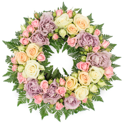 Centred on a clean white background, a lush circular wreath of roses and greenery forms a continuous halo of colour and texture, ideal as a funeral or memorial tribute. The eye first catches the velvety spiral of the roses: large, open blooms in ivory cream, gentle peach, and pale buttery yellow are interwoven with distinctive dusty lavender and mauve roses that lend an almost heirloom, antique feeling to the design. Between the bigger flowers, small clusters of blush pink spray roses, some in tight bud and others half-open, add intricate detail, while occasional pale green buds hint at the softness of new petals still hidden. The flowers sit on a base of rich, dark green fern fronds, their finely divided leaves emerging from between the blooms to create a deep, verdant backdrop that frames the lighter tones. The lighting is even and natural, highlighting the layered petals and the subtle gradations of colour without glare or harsh shadow. The ring itself is perfectly round, symbolising eternity and the continuity of memory, making it a fitting choice for families in Golders Green marking a farewell at local chapels or cemeteries. The overall mood is one of quiet peace and gentle consolation, like a softly spoken goodbye expressed through fresh, carefully placed blooms that look as though they were arranged that morning by an experienced local florist who understands the sensitivity of such moments.