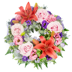 Centred on a crisp white background, this sympathy wreath forms a perfect circle of colour and texture, the kind of tribute often chosen for a funeral or memorial in Golders Green's calm residential streets. The arrangement is densely packed so that no gaps show, just a continuous flow of flowers and greenery. Soft, blush-pink and lavender-pink roses are scattered all around the ring, their velvety petals fully open and gently spiralling toward the centre of each bloom, giving an impression of tenderness and care. Intertwined with them are lisianthus blossoms in complementary shades: delicate pale pinks mingling with richer purple tones, their ruffled petals adding depth and a gentle, almost feathery softness. Two large coral-orange Asiatic lilies, one near the top of the wreath and another slightly off-centre near the bottom, draw the eye with their open, star-like petals and visible stamens, bringing a warm, hopeful accent to the cooler shades. Several unopened lily buds are nestled nearby, suggesting the quiet promise of blooms yet to come. The floral ring is anchored by abundant foliage, including rounded silvery eucalyptus leaves, glossy green stems, and fine fern fronds that give a flowing, natural outline. Blue-grey eryngium thistles punctuate the greenery with their spiky, cone-shaped centres, adding a striking, contemporary detail. Wisps of lavender-blue filler flowers, likely statice, weave between the larger blooms like a light haze. Near the lower section, folded bright green leaves are artfully shaped, a sign of skilled, modern floristry you might expect from a local shop close to Golders Green station. Together, these elements create a wreath that feels both respectful and uplifting, a graceful expression of sympathy suitable for a chapel service or a farewell gathering near Golders Hill Park.