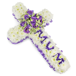 This carefully composed memorial cross is formed from hundreds of small white chrysanthemums, each bloom positioned to produce a seamless, cloud-like field of white that reads as purity and calm. A pleated ribbon of ruffled purple satin traces the entire outline, its subtle sheen and deep hue creating an elegant border that separates the white heart of the cross from its clean background. The vertical arm bears a personal inscription-MUM-crafted in bold, well-defined letters using clusters of dark purple eustoma or spray chrysanthemum heads; those purple letters sit in gentle relief against the white base and convey a direct, intimate message of love. At the cross's centre a lush spray gathers richly coloured elements: deep violet roses blended with lighter lavender roses and ruffled lisianthus, their textures moving from tight buds to softer, open cups. Variegated greenery with pale margins and darker centres nestles through the cluster, and small green berries add tactile contrast and depth.

The composition feels balanced, solemn, and consoling-its cool purple palette invites reflection while the soft petal density suggests tenderness. There is a faint floral, powdery scent and a crafted weight to the piece that comforts at a farewell. Flowers Golders Green prepares tributes like this for services at Golders Green Crematorium, for wakes near Golders Green Station, and for same-day delivery across the local area when families need a thoughtful, handcrafted expression of sympathy.