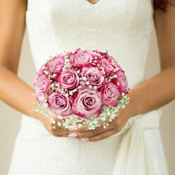 Imagine a Golders Green bride on her wedding morning, gently clasping a hand-tied bouquet that radiates both devotion and artistry. A dozen lush, two-tone pink roses bloom in the center, their soft, creamy cores melting into vibrant fuchsia outer petals, each one perfectly sculpted and suggestive of youthful hope. Wisps of baby's breath, like the first Chiltern air of a new day, weave between the roses, imparting a cloudlike softness and purity that contrasts beautifully with the saturated tones. The bride's hands-warm, lightly tanned, and elegantly manicured-hold the bouquet with quiet anticipation just above the intricate lacework of her white dress's bodice and delicate V neckline. The flowing fabric of a pale sash adds gentle movement, echoing the understated glamour of the ceremony. All around, the background is gently blurred, drawing the focus entirely to the intimate details of the flowers and the bride's poised embrace. This bouquet, crafted for lasting memories along Finchley Road and beyond, embodies the very heart of romance and commitment, evoking the peaceful yet joyous promise unique to weddings celebrated in Golders Green.