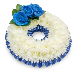 Captured with a soft overhead light that throws a delicate shadow beneath it, this circular memorial wreath reads as a composed, elegant statement of remembrance tailored for services in Golders Green and the surrounding neighbourhoods. The dominant form is a dense band of pure white chrysanthemums, hundreds of tiny, feathery petals layered so closely they form a soft, pillowed texture, their centers carrying the faintest wash of creamy yellow warmth beneath the cool white. Nestled on the upper-left curve are three deep cobalt-blue roses, their velveteen petals spiralling inward with a refined, almost velvet-like depth; tucked among dark green leaves, they create a poignant focal point against the snow of chrysanthemums. Framing both the inner and outer circumferences is a ruffled satin ribbon in matching deep blue, pleated neatly to catch highlights and provide a formal, finished edge that unifies the design. The subtle sheen of the ribbon, the tactile contrast between soft petals and crisp foliage, and the wreath's measured scale make it suitable for placement at Golders Green Crematorium or a local church doorway. At Flowers Golders Green we often craft such pieces when families request a tribute that balances tradition and personal colour; the palette suggests purity and grief softened by a dignified, calming blue. The wreath's understated scent and the quiet geometry of its construction are intended to console and focus attention on the memory it honours.