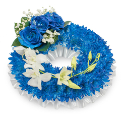 A lovingly constructed memorial wreath photographed against a pure white background, its language of colour and texture intended to convey comfort and dignity to mourners in Golders Green and beyond. The foundation is an abundant circle of vivid, deep-blue chrysanthemums-countless small heads packed closely so the surface reads as a lush, textural cushion of royal blue. Beneath and framing that blue is a pleated white satin ribbon, soft and glossy, which gives the design a quiet architectural edge and a refined, understated contrast. Near the upper-left quadrant three full blue roses form a focal cluster; their velvety, azure petals open in generous spirals, surrounded by tiny white gypsophila sprigs and a few dark-green leaves that add classic sympathy floristry detail. A sweep of pristine white orchids-Phalaenopsis in appearance-cascades from mid-left toward the lower-right, their smooth, sculpted petals and pale centres creating an airy, contemplative line across the blue field. Interspersed among these orchids are several fresh green buds, some tight, some just unfurling, suggesting hope and continuity amid remembrance. The arrangement carries varied textures-spiky chrysanthemum tips, soft rose velvet, silky orchid petals-and a faint, clean floral scent, finished with professional wiring and pleating techniques we use for local orders. As prepared by Flowers Golders Green for families arranging services near Golders Green Crematorium, Finchley Road or Temple Fortune, this wreath reads as a dignified, sympathetic tribute that feels both contemporary and timeless.