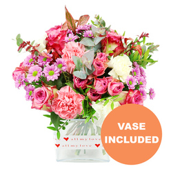 A richly detailed romantic bouquet fills the frame, arranged with the care of an experienced local florist familiar with Golders Green's cosy homes and leafy streets. The flowers rise from a clear, square glass vase, set against a pristine white background that accentuates every tone of pink, purple, and green. Several large roses in shades of fuchsia and soft blush pink form the focal point, their petals layered and slightly curled at the tips, some brushed with deeper magenta that adds drama and depth. Nestled beside them are ample carnations in warm coral and peach-pink, their ruffled petals creating a soft, almost feathery texture that feels both generous and comforting, like a hug sent in floral form. Cheerful lavender and purple daisy-like chrysanthemums with electric green centres pop up throughout the design, bringing a sense of movement and playfulness, as though they were picked on a bright morning stroll towards Golders Green station. The bouquet is wrapped visually in lush greenery: rounded eucalyptus leaves in silvery green, glossy darker foliage, and subtle touches of reddish-brown leaves that lend a hint of seasonal romance. The vase itself, with its clean lines and transparent sides, is decorated around the base by a white band sprinkled with vivid red hearts and the handwritten-style phrase "all my love," quietly reinforcing the sentimental message. In the lower right corner of the image, an orange circular badge announces "VASE INCLUDED" in bold white letters, signalling that this is a complete, ready-to-display gift. The arrangement feels ideal for birthdays, anniversaries, or a surprise same-day delivery around Temple Fortune or Hampstead Garden Suburb, radiating warmth, affection, and modern elegance.