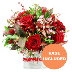 A luxurious dome-shaped bouquet filled with rich red blooms is photographed against a pure white background, as if displayed in the window of a Golders Green florist on a bright morning. Dominating the composition are velvety red roses, some in tight buds promising days of beauty, others fully unfurled to reveal their layered petals and evocative depth of colour. Threaded between them are clusters of deep crimson flowers resembling alstroemeria, their multiple petals curling and folding to create an intricate counterpoint to the classic roses. Interspersed throughout the arrangement, tiny sprays of pale pink and white filler blooms, akin to waxflowers, weave a light, cloud-like texture that gently lifts the heavier tones. Encircling this heart of crimson is a lush array of greenery: glossy dark leaves, softer green fronds, and distinctive burgundy and reddish-brown foliage, some with elegant veining that catches the light. These darker leaves lend a sophisticated, almost dramatic frame, much like the quiet, tree-lined streets just off Finchley Road framing the bustle of Golders Green. The bouquet is carefully placed in a clear rectangular glass vase with softly rounded edges, its transparency allowing a glimpse of the green stems and water, adding a sense of freshness and modern simplicity suited to contemporary flats and townhouses. A vivid red ribbon decorated with small white heart outlines is wrapped around the vase, subtly suggesting romance and affection without feeling overly ornate. In the lower right corner, a circular orange badge stands out, carrying the bold white text "VASE INCLUDED," instantly signalling that this arrangement arrives ready to display. The overall mood is one of passionate elegance, ideal for celebrating an anniversary, surprising a partner after a stroll through Golders Hill Park, or sending a heartfelt romantic gesture across Golders Green.