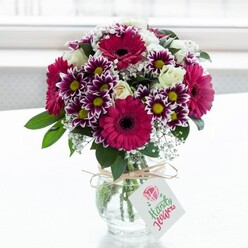 In this image, a richly coloured, professionally arranged bouquet sits centre stage in a sturdy, clear glass vase, the stems neatly gathered and visible through the slightly textured surface. The flowers form a joyful dome of colour: striking deep fuchsia and magenta gerbera daisies with dark, velvety centres draw the eye first, framed by generous clusters of bicolour chrysanthemums whose feathery petals are painted in shades of royal purple fading into clean white, each bloom circling a sunny yellow heart. Threaded between these bold flowers are creamy white roses and spray roses, some fully unfurled and others still half-closed, suggesting freshness and the promise of days of enjoyment. Wisps of white baby's breath drift among the larger blooms like tiny stars, lending lightness and a soft, romantic haze, while rich, deep green foliage provides structure and contrast, grounding the design. Around the neck of the low, jar-style vase, a length of rustic twine is tied into a casual yet deliberate bow, from which hangs a small rectangular tag marked with a pink rose illustration and fine script, hinting at a handwritten message from someone nearby in Golders Green. The background is a bright, softly diffused white, reminiscent of a sunny morning by a North End Road window, so the colours appear especially vivid and fresh. The arrangement feels like a celebration caught in full bloom - a bouquet suited to birthdays, congratulations, or a surprise delivery to a loved one in the NW11 area, bringing a burst of colour and happiness into any Golders Green home.