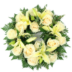 Seen directly from above against a clean white surface, this sympathy wreath is a study in understated elegance: creamy white roses and pale yellow lilies are arranged in a dense, perfectly circular composition set into a lush foundation of varied greenery. Approximately a dozen large, fully opened roses with their classic, spiralling petals create a soft, ivory focal band. Interspersed with them, graceful trumpet lilies in a tender lemon hue open in arcs or sit poised as buds, offering a contrast of scale and promise of future bloom. The greens are layered and textural - delicate fern fronds furnish a feathery fringe while clusters of rounded, blue-green leaves, suggestive of eucalyptus, supply depth and a muted contrast to the light blooms. The lighting is gentle and even, accentuating the creamy tones and casting minimal shadow, so the wreath reads as calm and luminous. There is an implied scent in the composition: a mild, sweet rose perfume tempered by the fresher, green-citrus hint of lilies. As a funeral tribute, the circular form and tranquil palette communicate continuity, respect, and solace. A local florist in Golders Green would select these particular stems to convey quiet dignity at a chapel service, graveside ceremony, or as a comforting memorial gift to a family - an arrangement that holds space for grief while offering a visual expression of remembrance and peace.