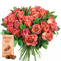 As someone who has crafted countless bouquets for the vibrant Golders Green community, I feel a particular fondness for this luxurious pairing of artfully arranged roses and gourmet chocolates. Nestled on a pure white background, the bouquet seems to glow with an inner warmth-roses in every stage of bloom, their petals transitioning fluidly from apricot and pale peach in the hearts, to salmon, dusky pink, and rich terra cotta on the outermost whorls. Subtle hints of yellow and cream breathe light into the arrangement, giving it an ethereal, late-afternoon quality, as if touched by the sun in Golders Hill Park. Intertwined through the roses, glossy green eucalyptus leaves provide both structure and aromatic freshness, framing the flowers like a jewelled garland. At the foot of this floral celebration, a Thorntons Orange Crisp chocolate pouch rests invitingly, displaying illustrations of decadent truffles with notes of orange zest and crisp wafer. Two unwrapped chocolates, glossy and deep brown, lie beside the packaging, promising an indulgence as sweet as a fond memory shared at the local cafés of Golders Green. This image calls to mind every reason we gather and give-a birthday toast, an anniversary dinner, a spontaneous gesture of gratitude-and echoes the generosity and warmth found throughout our neighbourhood. It is a composition that delights all senses, brimming with colour, fragrance, and heartfelt intention.