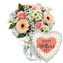 Set against a crisp white background, this Mother's Day scene feels like a quiet moment in a Golders Green flower studio, where every stem has been placed with care. A clear glass vase holds a generously rounded bouquet, its neck cinched with a simple loop of natural twine that gives the arrangement a relaxed, homespun charm. The flowers themselves glow in soft pastel tones: peach-pink roses open wide to reveal elegantly layered spirals of petals, their gentle blush colour evoking calm weekend walks through the gardens of Golders Hill Park. Nestled beside them, bold peach gerbera daisies, each with a deep chocolate-brown centre, introduce a joyful, sunlit energy. Between these focal blooms, airy sprays of white gypsophila drift like tiny clouds, while petite white chrysanthemums with subtle apple-green centres build a cushion of texture and volume, recalling small cottage flowers gathered from local gardens off Finchley Road. Fresh green foliage threads throughout the design, adding contrast and framing the lighter blooms with a leafy, natural border. Just behind the flowers, almost hidden, sits a small white card with hints of pink and green writing, suggesting a personal Mother's Day dedication. In front and slightly to the right of the vase, a glossy heart-shaped foil balloon becomes a playful companion to the bouquet. The balloon's white surface is dotted with glimmering gold and silver confetti spots, and at its centre lies a peach heart carrying the message "HAPPY Mother's DAY" in bold black type and elegant script. Together, the flowers and balloon form a warm, celebratory display, perfect for a Mother's Day delivery across Golders Green and the surrounding neighbourhood.