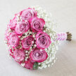 Set against a softly textured, light beige background that recalls a calm, linen-draped table in a Golders Green townhouse, this bouquet appears as a carefully composed love story. A cluster of sumptuous roses forms the focal point, each bloom moving from a gentle, dusty lavender-pink at its centre to a vivid fuchsia and deep magenta at the outermost edges. The petals curl inward in tight, graceful spirals or open just enough to show their sculpted layers, giving the arrangement a sense of depth and movement. Encircling the roses, a generous spray of white baby's breath creates a delicate mist, its countless tiny flowers weaving through the larger heads like scattered stars. Dark green foliage is tucked discreetly beneath the blossoms, providing structure and a fresh garden feel, as if the bouquet were gathered from a well-tended North West London garden. The stems are bound neatly at the base with a smooth lilac satin ribbon, its sheen catching ambient light. Twined around the ribbon is a refined string of small, white pearls, lending the design the quiet luxury so often chosen for weddings and significant celebrations. The bouquet feels as if it is waiting to be lifted by a bride stepping out of a Golders Green synagogue or registry office, or carried to an intimate anniversary dinner nearby. There is a mood of poised romance and thoughtful craftsmanship that suggests local florist expertise, where every bloom is placed to balance colour, texture, and sentiment perfectly.