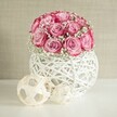 An elegant indoor still life features a domed bouquet of soft pink roses and airy white baby's breath set in a sculptural white vase, styled as if ready for delivery to a calm Golders Green sitting room. Around fifteen to twenty roses form a perfect rounded canopy, each bloom showing a gentle ombré from pale blush at the heart to deeper, almost fuchsia pink along the outer petals. The roses look freshly opened, with velvety petals that suggest a light, sweet fragrance, while clusters of tiny white gypsophila weave between them like delicate clouds, softening the overall shape. The bouquet rises from a spherical white container crafted from interwoven strands that resemble painted wicker or rattan, its open, criss-cross pattern letting light pass through and cast subtle shadows on the pristine white surface beneath. To the left, two decorative spheres made from slender, pale wooden strips echo the vase's woven texture, one larger and one smaller, adding a relaxed, natural accent reminiscent of the calm you might feel walking through nearby Golders Hill Park. The background is a warm, gently textured beige wall that recedes quietly, allowing the pink and white tones to glow under soft, even lighting. The composition feels serene and contemporary yet romantic, perfect for a Golders Green home office shelf or dining table, and ideal for occasions like anniversaries, Mother's Day, or a simple thinking of you gesture. It captures the atmosphere of a bright spring morning indoors, with every detail focused on texture, balance, and understated charm.