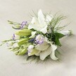 A lushly composed wedding bouquet lies horizontally upon a pale linen cloth, its arrangement speaking of both classic elegance and the gentle romance found in Golders Green. Large, fragrant white Oriental lilies command the focus, their trumpet blooms wide open to reveal delicate speckles and luminous yellow-green stamens, while others begin to unfurl, promising further beauty. Interlaced among these majestic lilies, clusters of white and deep purple lisianthus offer a graceful nod to twilight petals seen in Golders Hill Park. Their ruffled petals, edged in vibrant violet, invite the eye to linger, as unopened buds rest nearby, full of anticipation for the day ahead. Sprigs of fine baby's breath scatter like faint morning mist, lending airiness, while wispy asparagus fern and glossy green leaves provide fullness, natural movement, and deep woodland aroma-echoing the tranquil gardens lining Golders Green streets. The bouquet's stems are neatly wrapped in a white ribbon, reinforcing its bridal purpose. Created for heartfelt events, whether a wedding in a local synagogue or a celebration with friends in NW11, this bouquet radiates serenity, freshness, and a joyful sense of new beginnings-all hallmarks of local floristry.