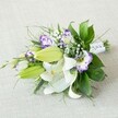 A hand-tied bouquet of white lilies, purple-edged lisianthus, and airy baby's breath rests diagonally across a softly textured light beige fabric, as if just placed on a linen-covered table in a calm Golders Green sitting room. The lilies are the stars of the arrangement, their broad, creamy-white petals fully open and gently curling back to reveal rich yellow-orange stamens dusted with pollen and a scattering of fine reddish speckles near the throat of each bloom. Around them, several elongated buds in fresh chartreuse green stretch outward, still closed and smooth, promising more blossoms in the days ahead. Threaded between the lilies, you can see clusters of Eustoma (lisianthus) in white, each ruffled petal tipped and washed with shades of lavender and deep violet, so the flowers appear like little swirling cups of purple and cream. Tiny white Gypsophila florets float above and between the larger flowers, like a soft mist weaving through the bouquet, adding lightness and a romantic haze. Lush foliage in several tones of green frames the blooms: some leaves are broad, glossy, and dark, catching the gentle, diffused light; others are finer and more linear, with wispy fronds and slightly spiky textures that create movement and depth. At the narrow end of the bouquet, the stems are neatly aligned and wrapped in a crisp white satin ribbon, subtly embellished with delicate bead-like details that suggest a bridal or special-occasion piece. The overall impression is of refined yet effortless elegance, a fragrant, fresh bouquet perfect for a wedding in Golders Green, a registry office celebration near Hampstead Garden Suburb, or a meaningful gift carried home from a trusted local florist after a peaceful walk through nearby Golders Hill Park.