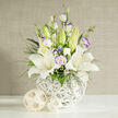 In this carefully composed image, a graceful white and lavender-toned bouquet appears as if ready to be delivered to a wedding venue near Golders Green Station. Dominating the arrangement are several open white lilies, their petals generous and silky, stretching outward in quiet confidence. The centers are speckled and gently shaded, with prominent yellow-orange stamens standing upright, suggesting a light, sweet fragrance. Nestled between these star-shaped blooms are ruffled lisianthus flowers, mostly in creamy white, with some petals kissed at the edges by soft lavender and pale violet, like a watercolour wash. Unopened lily buds in a cool, pale green hint at future blossoms, mirroring the promise of new beginnings celebrated in local synagogues and event spaces off Golders Green Road. Fine sprays of deep green foliage, along with lacy clusters of tiny white filler blooms, soften the gaps and bring texture, while a few deeper blue and purple florets introduce a subtle, refreshing contrast within the predominantly white design. The bouquet is cradled in a bulbous white vase made from intricately woven strands, its sculptural form reminiscent of a contemporary basket you might spot in a boutique along nearby Temple Fortune. This vase rests on a pristine white tabletop, enhancing the composition's purity and minimalist elegance. To the side, two decorative woven spheres in light natural tones echo the vase's pattern and add earthy warmth. The background is a muted beige with a quiet, vertical woven texture, ensuring the flowers remain the star. Overall, the photograph conveys calm sophistication, ideal for illustrating modern wedding flowers, thoughtful condolence arrangements, or refined interior styling in and around Golders Green.