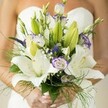Captured in the delicate morning light of Golders Green, this bridal bouquet conveys a serene harmony of colours and textures, lovingly arranged for a bride immersed in her special day. The composition is anchored by the luminous purity of white Oriental lilies, their petals wide and inviting, holding soft yellow stamens and marked with subtle, endearing speckles. Graceful lisianthus unfold around them, some blooms fully open to reveal petals edged in a whimsical lavender purple, others still curled in promise. Wispy sprigs of white baby's breath create a sense of ethereal movement and airiness, reminiscent of the gentle breezes common in local gardens and parks. Deep purple veronica blossoms bring a surprising contrast, their shape and shade reminiscent of the vibrant blooms found in community spaces like Golders Hill Park. Lush green foliage-ranging from broad leaves to feathery asparagus fern-spills naturally from the bouquet, overflowing with freshness and vitality. The bride's lightly polished nails softly frame the bouquet, grounding the moment in human warmth. Against the gentle blur of her white dress and bare shoulder, the florals become the day's true centrepiece, capturing the hopeful spirit and romantic anticipation cherished by couples throughout Golders Green. This handcrafted bouquet expresses both artistry and emotion, perfect for wedding ceremonies or bridal portraits set amidst the area's unique blend of elegance and neighbourhood charm.