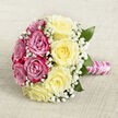 Resting on a softly woven, oatmeal-toned fabric, this classic hand-tied bouquet is captured from a slightly elevated angle that shows off its perfect round shape. The roses are arranged almost like a yin and yang of colour: one side an intense cluster of fuchsia and magenta-pink blooms, the other a serene sweep of creamy, pale yellow roses. Each blossom is in peak condition, petals unfurling in flawless spirals that suggest both richness and tenderness. Fine sprays of white baby's breath are woven through the entire arrangement, creating a misty halo effect that lightens the overall look and gives a gentle, bridal air you might associate with a ceremony at a Golders Green venue or a reception close to Brent Cross. Between the roses, small, glossy green leaves offer contrast and depth, making the colours seem even more vivid without overpowering the softness of the design. The stems are gathered cleanly into a smooth handle, bound in lustrous pink satin ribbon that feels silky and cool to the touch. Over the ribbon, a slim garland of white pearls is wound in a graceful spiral, like jewellery chosen to match a bride's dress. The neutral backdrop, with its subtle linen-style texture, keeps all the focus on the flowers and hints at a relaxed yet elegant setting, perhaps a local family celebration after a ceremony in nearby temples or parks. This bouquet's gentle fragrance and romantic two-tone palette make it ideal for weddings, vow renewals, or a significant anniversary, especially for couples celebrating their story in and around Golders Green.
