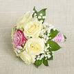 On a softly textured, light beige background that resembles fine linen or lightly woven burlap, a small hand-crafted bouquet is displayed at a gentle diagonal, as though just set down between moments of celebration. Five creamy white roses form the core of the design, their petals unfurling in concentric layers that feel plush and velvety, like the quiet luxury of a classic North West London wedding. Just to the front-left, a single vivid fuchsia-pink rose breaks the calm palette with a joyful splash of colour, immediately capturing attention without overpowering the surrounding ivory blooms. Delicate clouds of baby's breath are woven throughout the bouquet, their tiny white flowers clustering like frost or soft mist, filling the gaps between roses and adding a dreamy, ethereal quality. Around the edges, deep green, tapering leaves, perhaps ruscus or salal, frame the flowers and lend a sense of fullness and gentle structure, the polished greens contrasting beautifully with the softer tones. The stems are bound tightly with a bright pink satin ribbon, smooth and luminous, shaped into a petite handle that feels perfect for a corsage, boutonniere, or a small bridal party bouquet. Over this ribbon, parallel rows of tiny white pearls have been meticulously placed, giving a refined, almost vintage elegance that would look at home at a wedding reception near Golders Green station or a prom at a nearby school. The lighting is soft and even, allowing every texture-the satin sheen, the rose petals, the airy baby's breath-to be seen clearly, and the overall impression is one of romance, care, and thoughtful craftsmanship suited to meaningful moments shared across the Golders Green community.