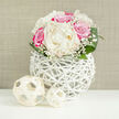 On a clean white surface, a compact, domed bouquet of white peonies and vivid fuchsia-pink roses draws the eye, like a small, radiant island of colour in a serene room. The peonies are at their lush peak: generous ivory blooms, petals layered in soft folds that look almost velvety, their rounded forms anchored firmly at the heart of the design. Around them, the pink roses provide a richer, more saturated note, each flower carefully spaced so its spiral centre can be admired, their edges catching the light with a gentle sheen. Threaded through the larger blooms are delicate sprigs of baby's breath, each tiny white flower forming a cloud of fine texture that makes the arrangement feel light and breathable rather than heavy. The blooms emerge from a striking white vase formed from interwoven strands that resemble painted wicker or rattan, the open latticework creating a sculptural, basket-like shape. The stems and inner shadows can be glimpsed within this airy structure, giving the impression of craftsmanship and intention rather than a simple container. In front of the vase lie two small decorative spheres, also woven and pale in colour, echoing the vase's texture and adding a sense of calm balance. Behind everything, a softly textured beige backdrop-similar to woven fabric-keeps the focus on the flowers while lending warmth to the scene. The lighting feels like gentle daylight filtered through a Golders Green window, steady and flattering, making this arrangement ideal for a wedding breakfast table in a local venue, an intimate engagement celebration near Hampstead Garden Suburb, or an elegant gift delivered to a loved one across NW11.
