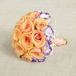 Framed on a neutral, lightly textured beige fabric, this bridal bouquet is an exercise in soft, romantic contrasts captured from a slightly raised viewpoint that reveals its rounded, abundant form. Peach and soft orange roses dominate the centre, each bloom opening in layered spirals that seem to cradle the air; their petals are satiny and hold a warm, sunlit glow. Interspersed among these classic shapes are crisp white lisianthus whose petals are kissed at the edges by a striking violet-purple - a contemporary twist that sharpens the arrangement's palette and draws the eye into each ruffled bloom. Wisps of baby's breath tuck into the gaps, lending the bouquet a whispering lightness against the roses' dense luxury. The stems are bound with a peach-coral satin ribbon, smooth to the touch, and a string of cream pearls winds around the handle in a deliberate, elegant coil, suggesting heirloom detail and bridal tradition. The composition rests on linen-like fabric that softens the scene and gives the photograph a tactile, intimate quality; the lighting is gentle and natural, flattering the warm tones without harsh shadows. As a florist attentive to North London sensibilities, I imagine delivering this to a wedding party near Finchley Road or setting it on a table at a tranquil reception in Childs Hill - a bouquet that combines classic charm with a modern edge and speaks softly of commitment, celebration, and the quiet joy of well-made things.