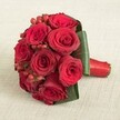 An exquisite, tightly clustered bouquet of ten sumptuous red roses is artfully displayed against a pale, softly textured background reminiscent of Golders Green's inviting spaces. Each rose is in peak bloom, with their velvety crimson petals perfectly unfurled, glowing softly under even, diffused daylight. Between the luscious blooms, small clusters of bright red hypericum berries add dimension and festive charm, catching the light and providing a lively pop of texture that conjures the spirit of celebrations in the heart of Golders Green. A halo of broad, glossy emerald leaves elegantly encircles the roses, framing the bouquet and adding structure, as if gently cradling each blossom. The stems are expertly gathered and wrapped with a rich, deep red satin ribbon, the polished finish of which offers a pleasing contrast in both texture and colour. The bouquet's classic, rounded shape and artful blend of rich reds and greens evoke deep affection, ideal for weddings, anniversaries, or romantic arrangements destined for delivery around Golders Green, Hampstead, or Childs Hill. This image beautifully conveys both the luxury of fresh florals and the heartfelt artistry of a local florist, inviting thoughts of cherished celebrations and timeless love blooming amidst Golders Green's leafy streets.