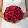 As if plucked from an exquisite Golders Green wedding, this evocative scene reveals a radiant bridal bouquet held close in the gentle embrace of a bride clad in a lace-adorned white dress. Lush, voluptuous red roses-some still shyly furled, others fully blossomed-conjure a sense of regal passion and enduring devotion. Sprinkled amid the blooms are clusters of lively hypericum berries, their bright red pops lending a cheerful playfulness that echoes the vibrancy of Golders Green's high street and local gardens. All are nestled within a collar of broad, glossy green leaves, offering elegant structure and a nod to the area's leafy parks. The bride's manicured hands cradle the bouquet, underscoring its importance, while soft, diffuse light highlights every velvet petal and glassy berry. This bouquet radiates warmth, tradition, and a gentle promise-an ideal image for those seeking inspiration for luxurious, locally inspired wedding flowers in Golders Green, where romance and tradition walk hand in hand through every season.