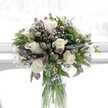 A softly lit, elegantly arranged bouquet of creamy white roses, lavender alstroemeria, and cloud-like baby's breath stands in a clear cylindrical glass vase, placed against a bright, gently diffused backdrop that feels like a sunlit Golders Green window. The roses are fully opened yet still delicate, their velvety petals spiralling outward in layers that suggest quiet romance and purity. Around them, clusters of alstroemeria in muted purples and lavender tones are shown in different stages of bloom, from slender closed buds to partially open trumpets streaked with fine, darker veining, adding subtle drama and texture. Wisps of white gypsophila weave between the larger blossoms, creating a light, misty halo that softens every edge. Deep, glossy green foliage forms a generous collar around the blooms, with broad leaves and slimmer lines of greenery providing structure and contrast, as if the flowers were freshly gathered from a well-tended garden near Golders Hill Park. Through the transparent vase, the neatly trimmed green stems are clearly visible, immersed in clean water that hints at just-cut freshness and expert care. The natural daylight falls gently across the arrangement, highlighting layers of white and lilac and casting the faintest shadows, giving the bouquet a serene and uplifting atmosphere. It feels like the kind of floral design someone in Golders Green might place on a hallway console after a special delivery, perfect for expressing sympathy, offering congratulations, or simply bringing a sense of calm beauty and new beginnings into a North West London home.