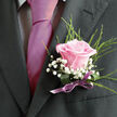 Pinned to a charcoal-grey suit, this wedding rose boutonniere appears almost like a painted detail brought to life, its soft pink bloom unfolding petal by petal with unhurried grace. The rose itself is a gentle, romantic shade-as if it has absorbed the blush of an early spring sunrise over Golders Hill Park-its velvety texture inviting the fingertips even though it rests perfectly still. Around the central flower, threads of fine greenery arc upward and outward, their slim, feathery needles framing the rose and giving the composition a fresh, slightly woodland feel. Nestled in between are tiny clusters of bright white gypsophila, their miniature blossoms catching the light like scattered pearls and lending an air of innocence that suits a wedding morning. The stems are carefully bound at the base with a smooth lilac satin ribbon, wrapped tight and tied in a tidy, symmetrical bow. The ribbon adds a touch of polish and a hint of playful colour that quietly echoes the tones of the groom's tie and pocket square. The overall mood is calm and anticipatory, as if captured just before stepping out of a townhouse near Golders Green Road for the walk to the ceremony. You can almost sense the faint, fresh fragrance of the rose mingling with the clean scent of pressed fabric, an intimate mix of floral sweetness and formality. Crafted with care and balance, this boutonniere becomes more than an accessory; it is a small emblem of affection and commitment, worn close to the heartbeat throughout the day.