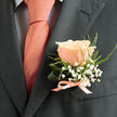 This boutonniere is an exercise in gentle contrast and whispered celebration, crafted to rest comfortably on a groom's lapel or a devoted guest's jacket. A singular peach rose commands the composition with soft, warm petals that spiral inward like the first light of dawn; its coral-pink tones are luminous without being loud. Surrounding the central bloom, delicate sprays of white baby's breath create a cloud-like cushion, their tiny blossoms scattering points of innocence and light. Fine green foliage-slender, slightly serrated leaves with a fresh, early-morning feel-frames the rose and lends structure while keeping the overall silhouette airy and elegant. The stems are bound with a light peach silk ribbon tied into a modest bow, the satin catching a subtle sheen that mirrors an accompanying tie.
Placed against a charcoal suit, the boutonniere's colours glow with added depth; the dark fabric makes the peach and white sing with clarity. Texture plays through the piece: the rose's velvet, the baby's breath's papery fineness, and the foliage's soft resilience invite touch without asking for it. A gentle floral perfume seems present, kind and hopeful-perfect for weddings, anniversaries, or vows exchanged near familiar Golders Green streets like Hoop Lane or Finchley Road. The arrangement is composed with the care of an experienced hand, balancing tradition and personality so that it reads as both ceremonious and affectionate. Lightweight, balanced, and quietly spirited, this boutonniere feels made for moments that matter.