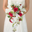 Held delicately at waist level, this cascading bridal bouquet becomes the centre of the scene, framed by the bride's arms and the subtle texture of her white lace gown. The composition begins with a rich, rounded heart of scarlet red roses, each bloom tightly packed and velvety, their deep tones suggesting passion and enduring love. Interwoven among them are open white lilies, their broad, star-like petals fanning outward and revealing green stamens that add a touch of fresh, natural colour. From this central cluster, a stream of white Phalaenopsis orchids flows downward, forming a soft waterfall effect; some blossoms are fully open, while others remain in bud, hinting at moments of beauty still to come. Wisps of baby's breath drift through the arrangement, like gentle clouds of white mist, softening the contrast between the red roses and crisp lilies. Bright red berries, reminiscent of hypericum, are nestled between the blooms and leaves, their round, glossy shapes catching the light and giving a sense of joyful celebration, ideal for a winter wedding in Golders Green or a chic civil ceremony near Temple Fortune. A mix of greenery, from airy fern-like fronds to broader, darker leaves, frames the flowers and extends the length of the teardrop design, guiding the eye downward in one fluid motion. The neutral, blurred background in warm beige tones ensures the bouquet and bridal silhouette remain the undisputed focus, allowing every nuance of colour and texture to stand out. The image feels like a quiet pause on a busy wedding morning, as if the bride has just stepped out from a salon on Golders Green Road, taking a breath before walking down the aisle with this elegant, handcrafted bouquet in her hands.